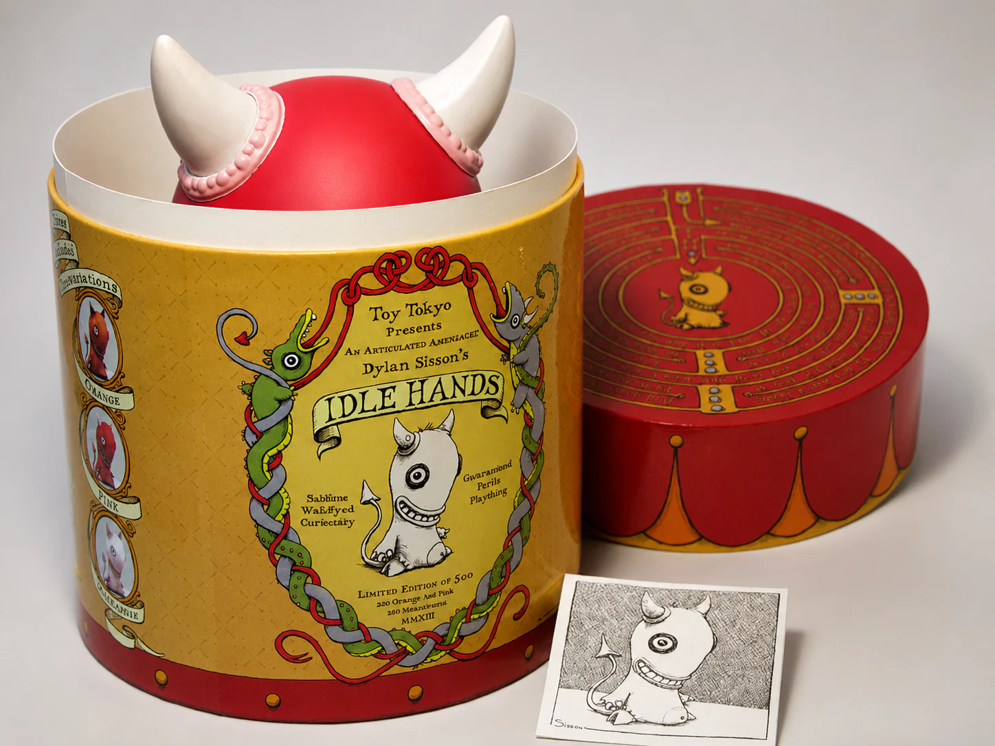 The pink Idle Hands figure peeking from its illustrated cylindrical package beside the maze lid and numbered coaster drawing.