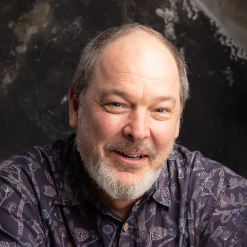 Dylan Sisson smiling in a square portrait against a dark textured background.