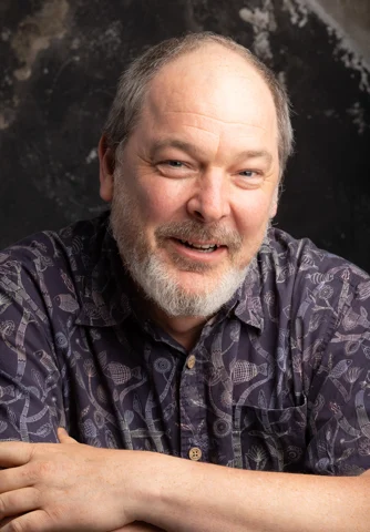 Dylan Sisson smiling with his arms resting in front of him, photographed vertically against a dark background.
