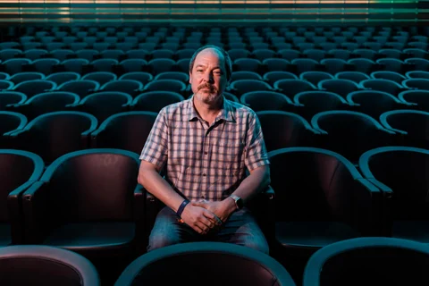 Dylan Sisson seated among rows of teal seats in a theater.