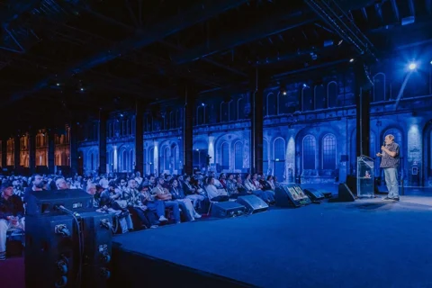 Dylan Sisson speaking onstage to a seated audience in a blue-lit auditorium.
