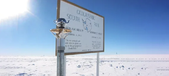 A silver Walking Teapot perched on the Geographic South Pole marker in bright Antarctic snow.